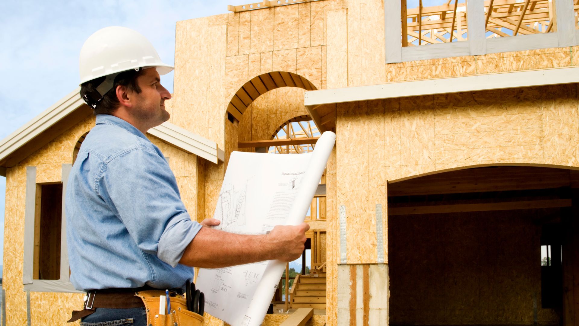 A man in a hard hat holding a blueprint in front of a house